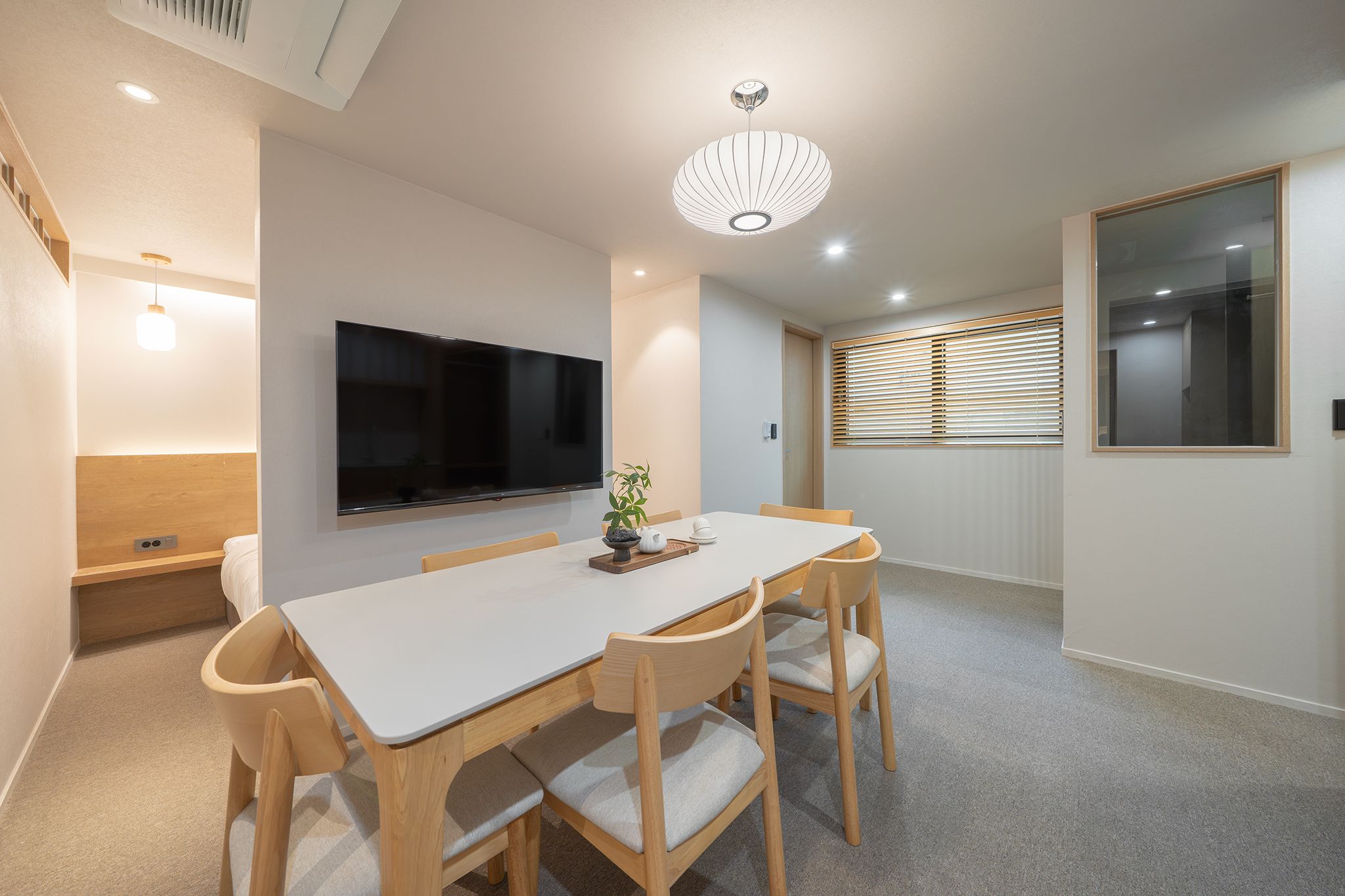 A modern dining area with a white table, wooden chairs, a flat-screen TV, and a ceiling light fixture.
