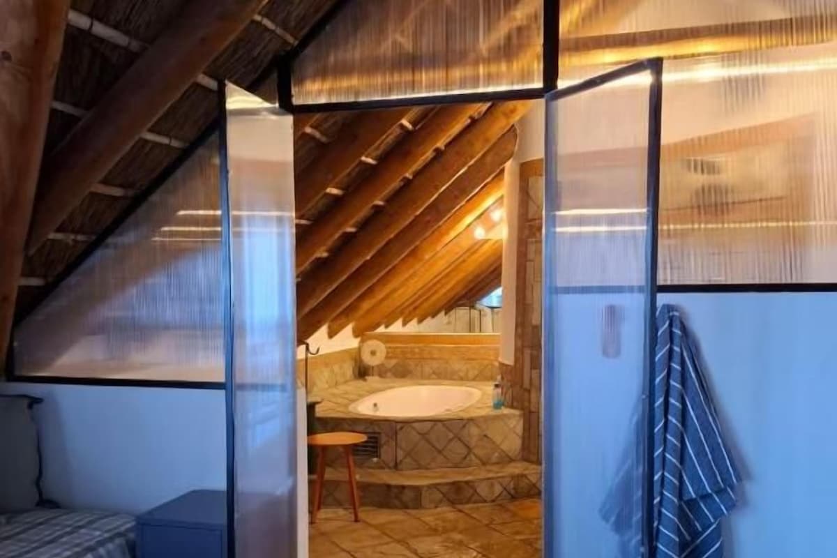 Un dormitorio con techo inclinado, una cama, una mesita pequeña y vistas a un baño con lavamanos y bañera.