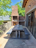 Un patio exterior con una parrilla de ladrillo, una mesa de madera con bancos y un ventanal que da a un área cubierta.