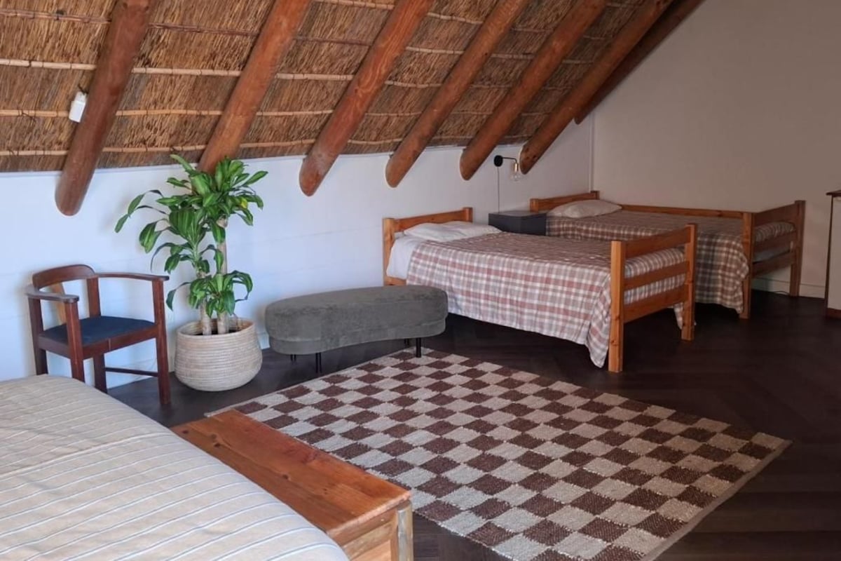 Una habitación con piso a cuadros, una cama, una silla, una mesita y una planta en maceta.