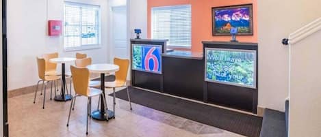 A hotel reception area with a counter, a TV screen displaying the number 6, and a seating area with chairs and tables.