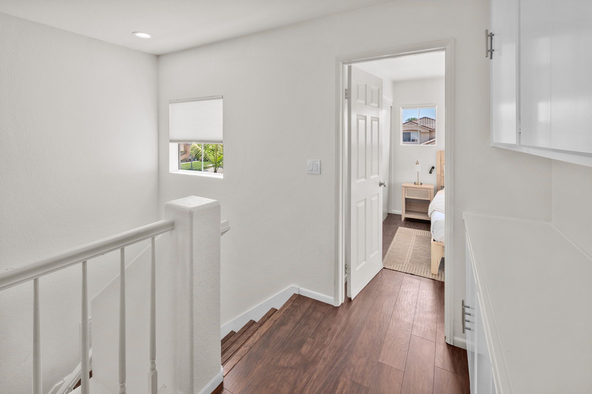 A hallway with a wooden floor, white walls, a window with a view, and a door leading to a bedroom.