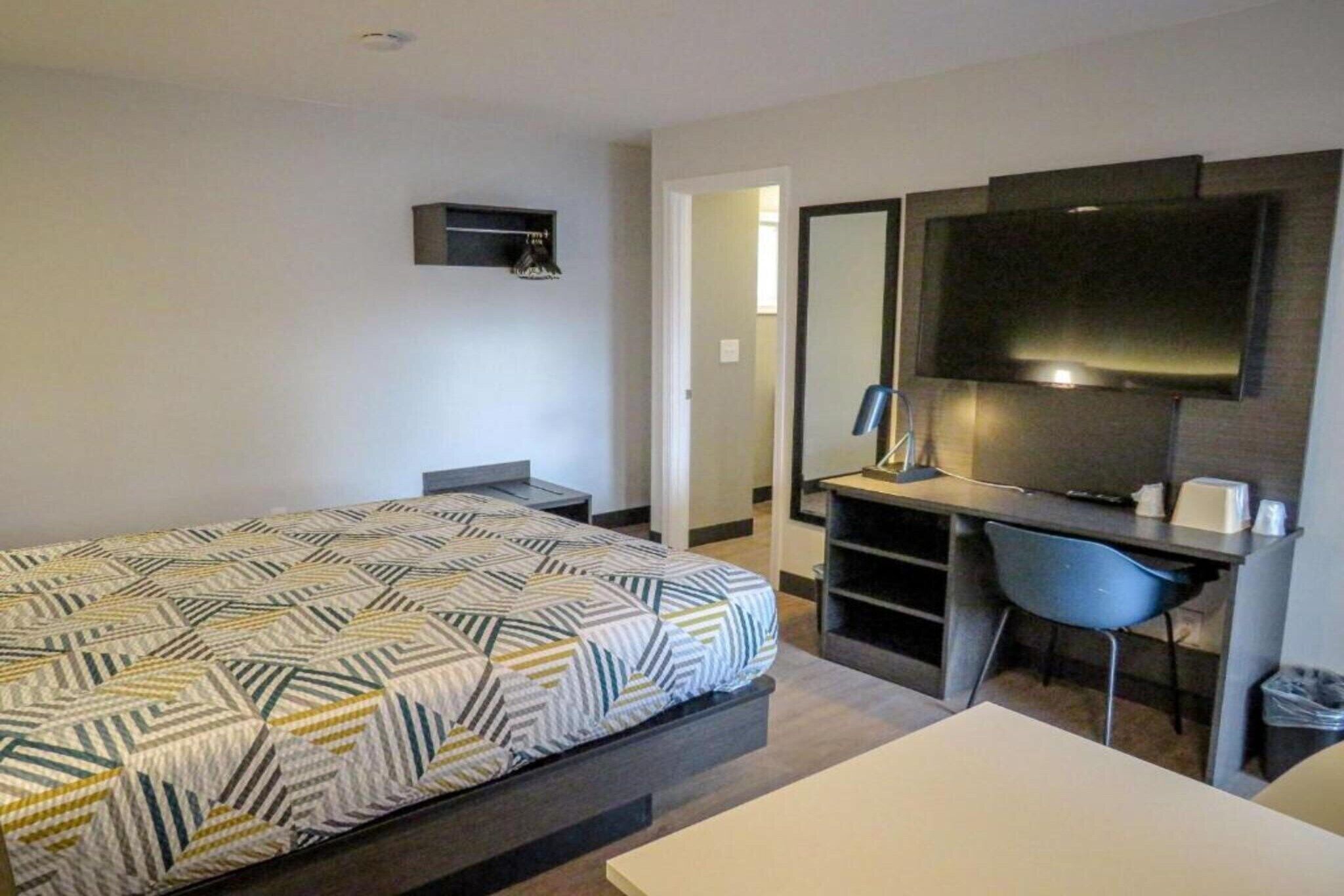Una habitación de hotel con cama, escritorio, silla, televisión y una pequeña estantería.