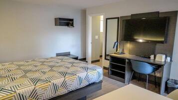 A hotel room with a bed, a desk, a chair, a TV, and a small shelf.