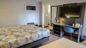 Una habitación de hotel con cama, escritorio, silla, televisión y una pequeña estantería.