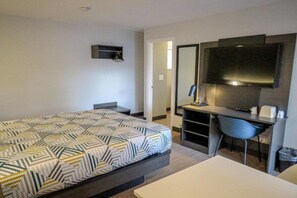 Ett hotellrum med en säng, ett skrivbord, en stol, en tv och en liten hylla.