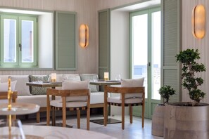 A room with a sofa, two wooden chairs, a small table, and a potted plant. - Blu Suites (Imerovigli)