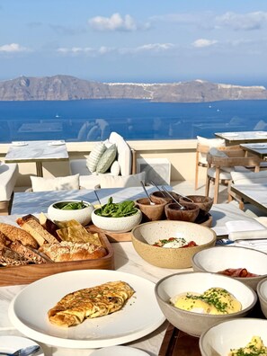 A table set with various dishes and bread, overlooking a scenic view of the sea and mountains. - Blu Suites (Imerovigli)