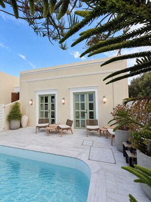 A modern outdoor pool area with a pool, lounge chairs, and a building with large windows and doors. - Blu Suites (Imerovigli)