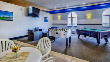 A room with a pool table, foosball table, and a table with chairs and a vase of flowers.