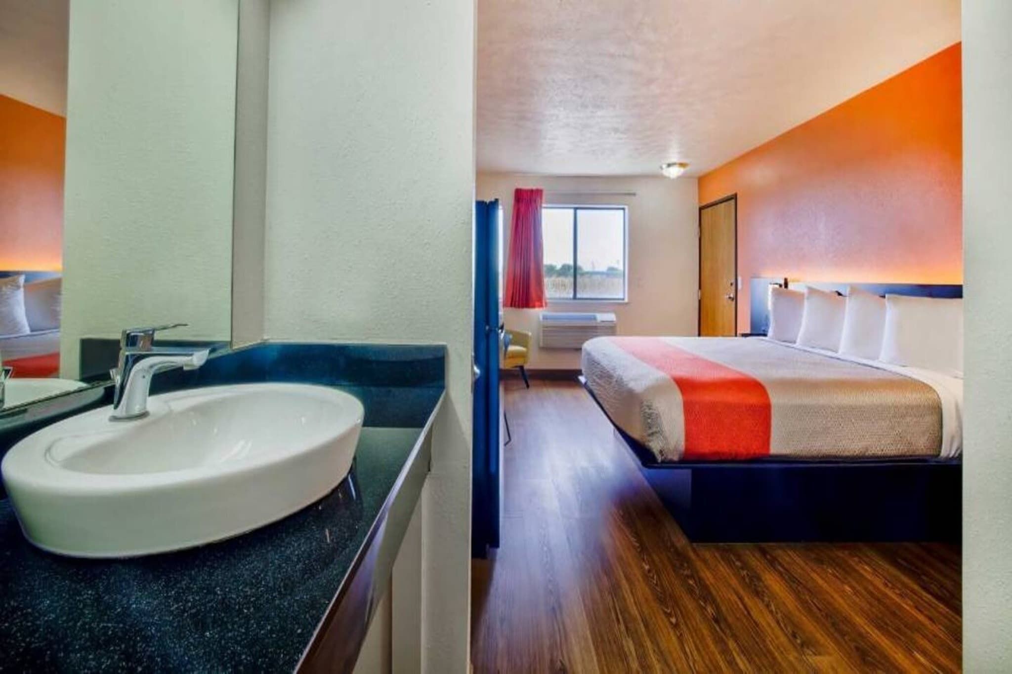 A hotel room with a large sink, a bed with a red accent, a window with curtains, and a wooden floor.
