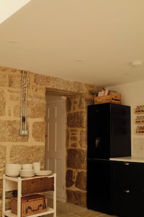 A room with a stone wall, a white door, a black cabinet, and a shelf with plates and baskets. - Artist's Cottage in Cornwall (Penzance)