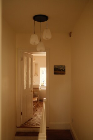 A hallway with a door leading to a bathroom, a small painting on the wall, and a light fixture above. - Artist's Cottage in Cornwall (Penzance)