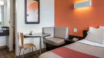 Een hotelkamer met een bed, een bureau, een stoel, een klein tafeltje, een nachtkastje en een wandlamp.