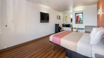 A hotel room with a bed, a desk, a chair, a television, and a kitchenette.