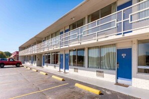 Un motel con una fila de habitaciones, una zona de estacionamiento techada y una camioneta roja estacionada afuera.