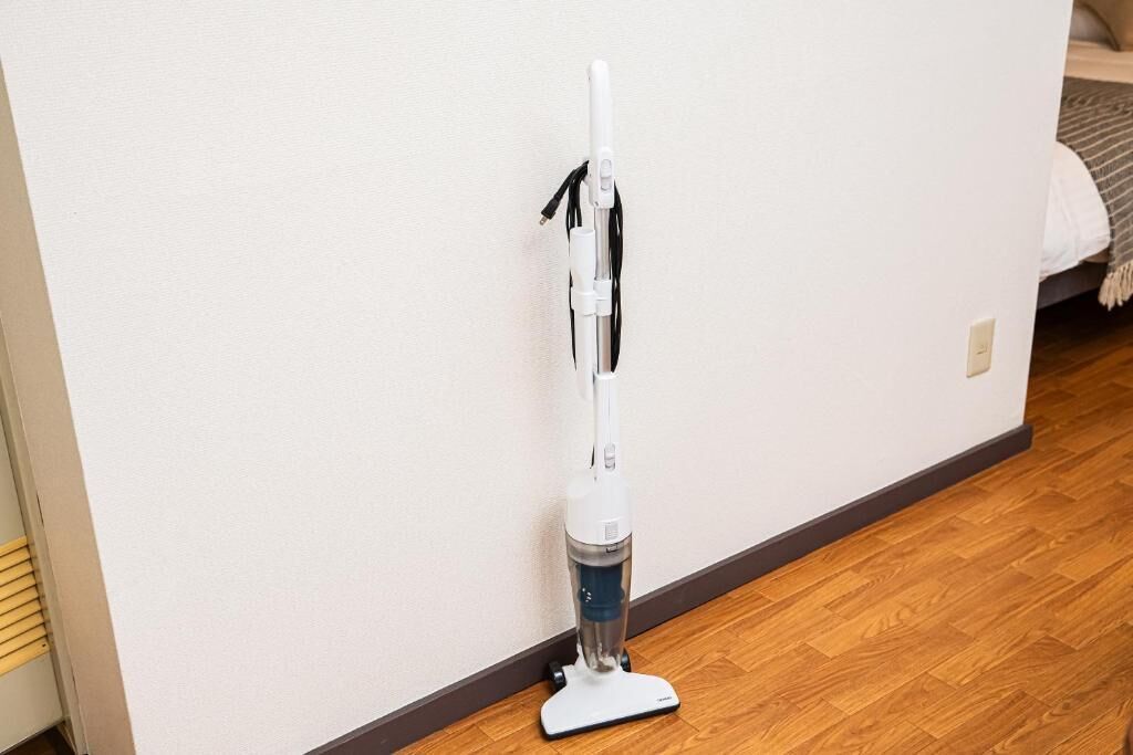 A white upright vacuum cleaner with a cord, placed against a wall with a wooden floor.
