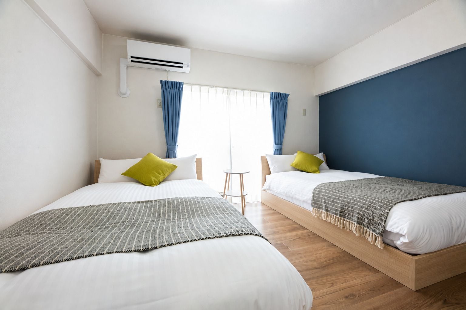 A hotel room with two beds, a small table, and an air conditioning unit.