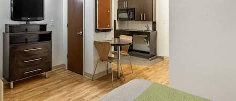 Een kleine kamer met een televisie, een bureau, een stoel en een kitchenette.