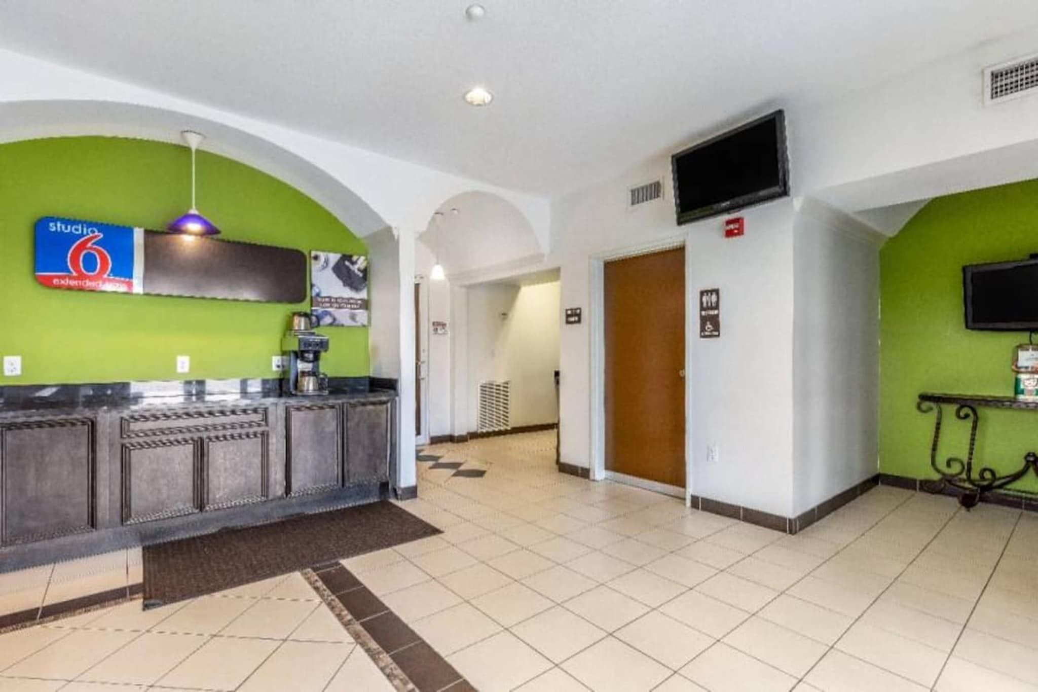 Um saguão de hotel com um balcão de recepção, uma máquina de caf é, uma televisão e uma placa indicando Studio 6.