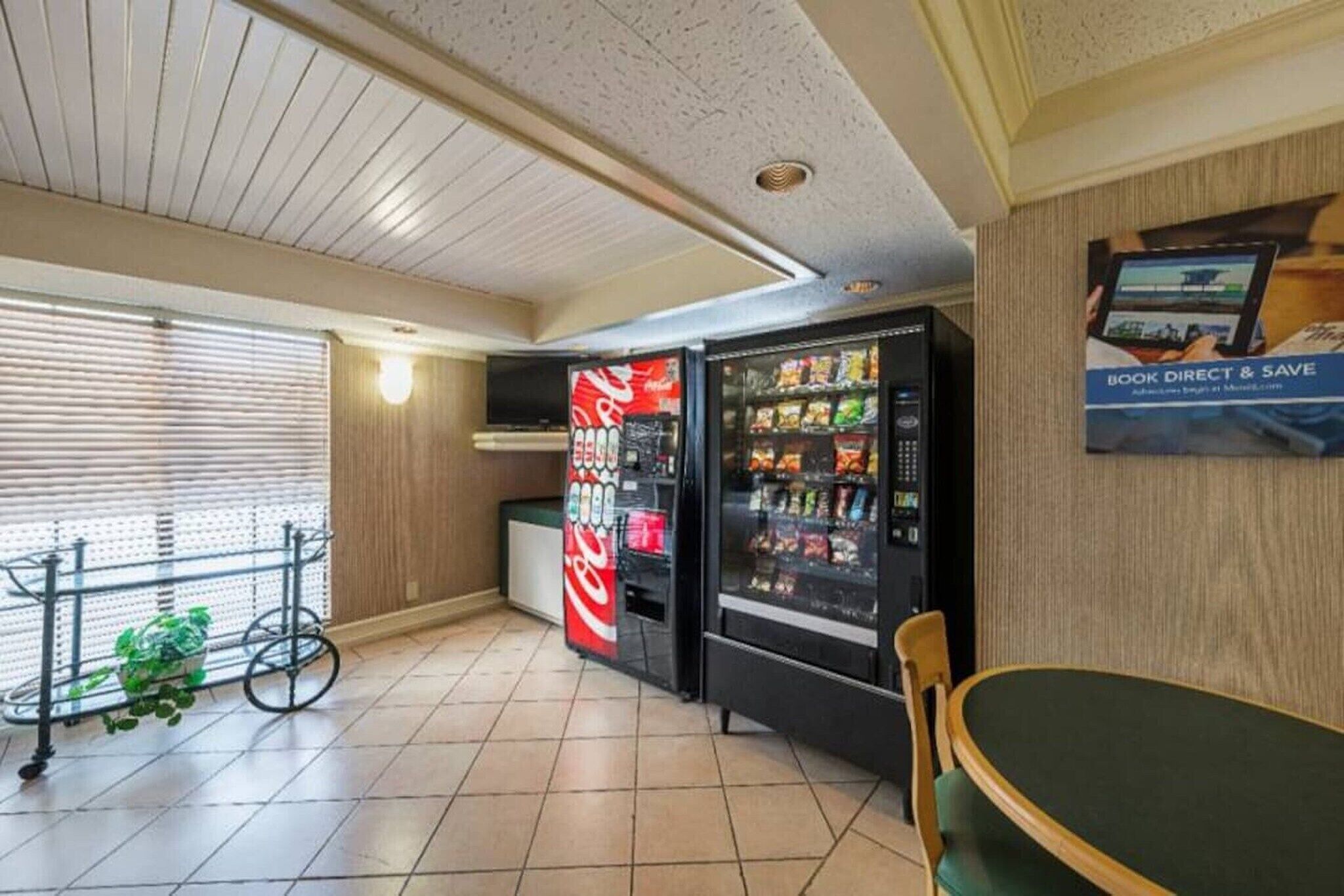 A hotel room with a dining table, two vending machines, a television, and a window with blinds.