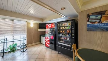 Ein Hotelzimmer mit Esstisch, zwei Getränkeautomaten, Fernseher und einem Fenster mit Jalousien.