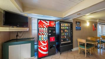 A room with a television, a Coca-Cola vending machine, a snack vending machine, and a wooden table with chairs.