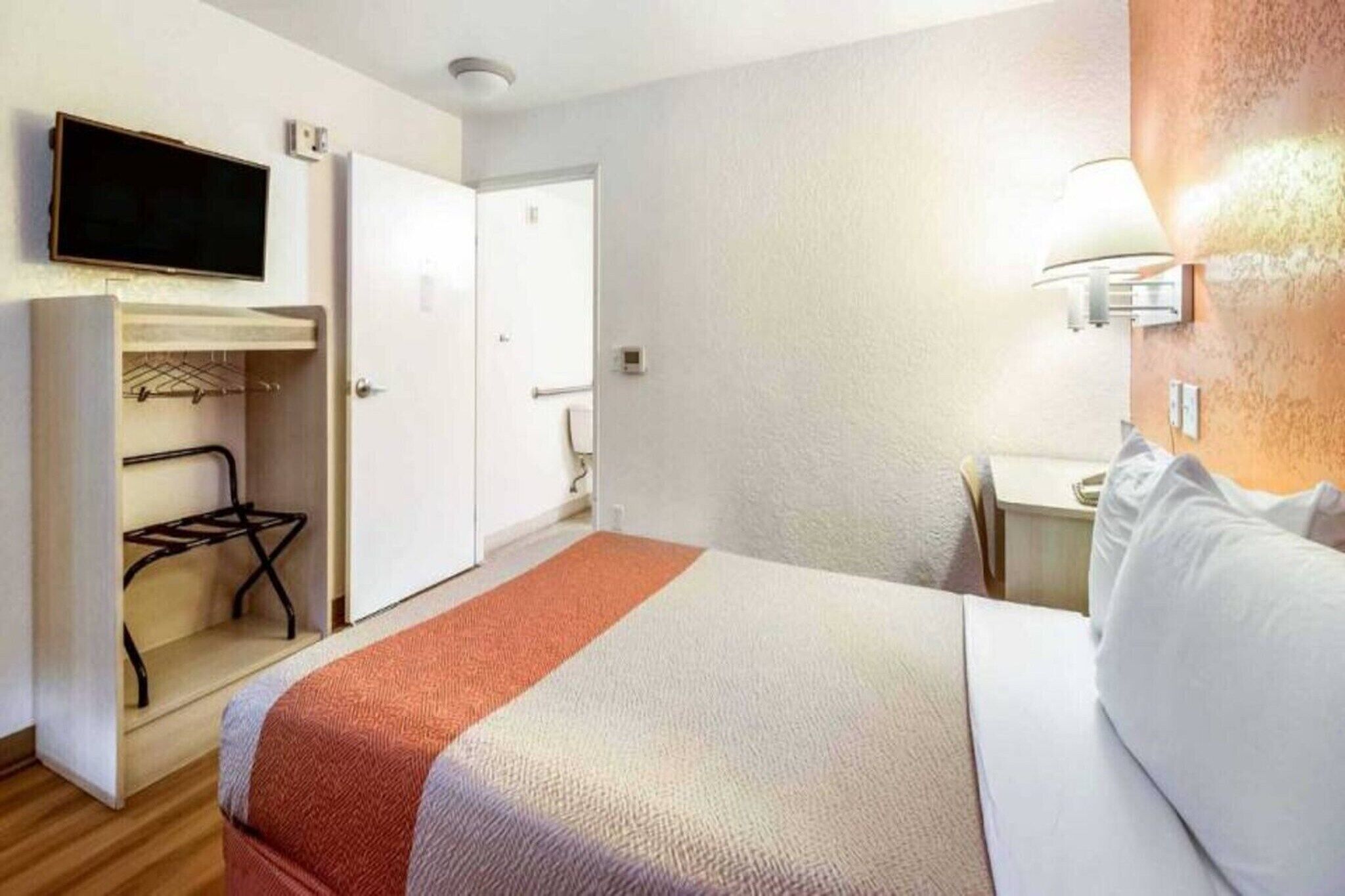 Ein Hotelzimmer mit einem Bett, einem an der Wand befestigten Fernseher, einem Kleiderschrank und einem Schreibtisch mit Lampe.
