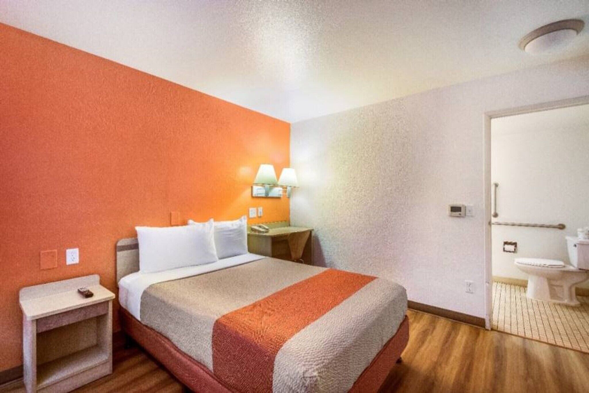 Habitación de hotel con cama, mesita de noche, lámpara, inodoro y zona de ducha.