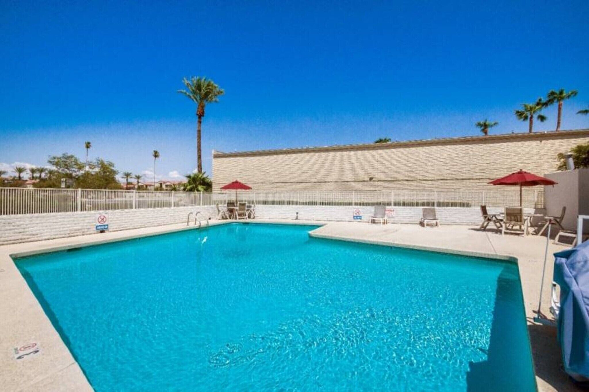 Piscina con sillas de descanso y sombrillas, rodeada por una valla blanca y palmeras.