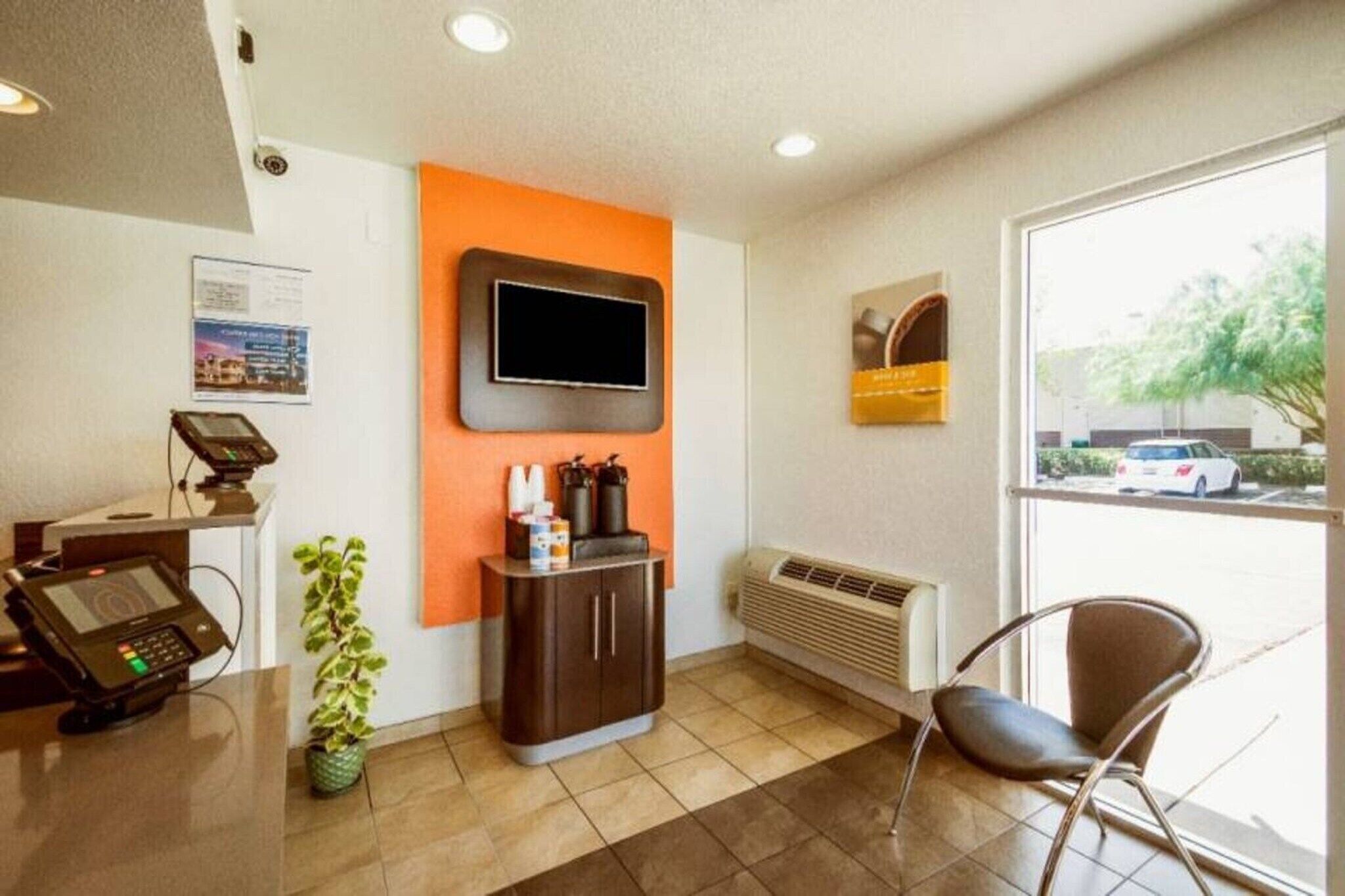 A reception area with a flat-screen TV mounted on the wall, a small cabinet with a plant, a chair, and a window with a view of a parking lot and trees.