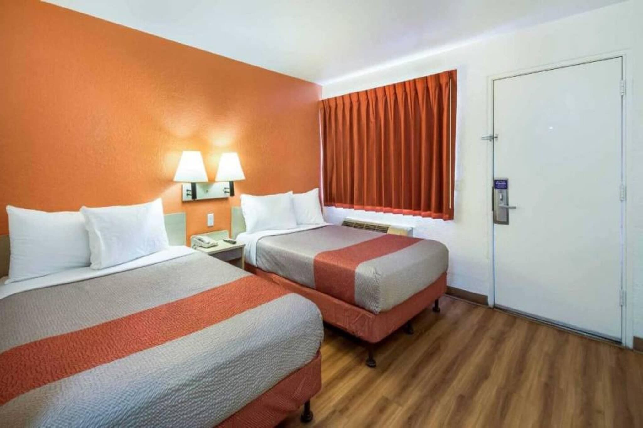 A hotel room with two beds, a nightstand, a lamp, a window with curtains, and a door.