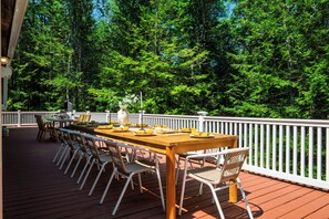 A wooden deck with a dining table set for six, surrounded by wicker chairs, overlooking a forested area.
