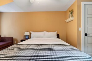 A hotel room with a bed, a nightstand with a lamp, a wall-mounted shelf with a plant, and a door.