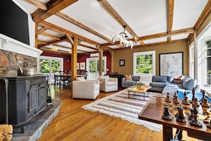 A spacious living room with a fireplace, wooden beams, a large window, and a chessboard.