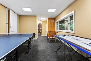 A room with ping pong tables, a foosball table, and a window with a view of trees.