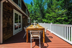 An outdoor dining area with a wooden deck, white railing, and a view of the surrounding forest.
