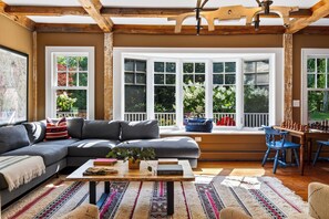A cozy living room with a sectional sofa, a coffee table with books and a plant, a chessboard, and a view of a garden through large windows.