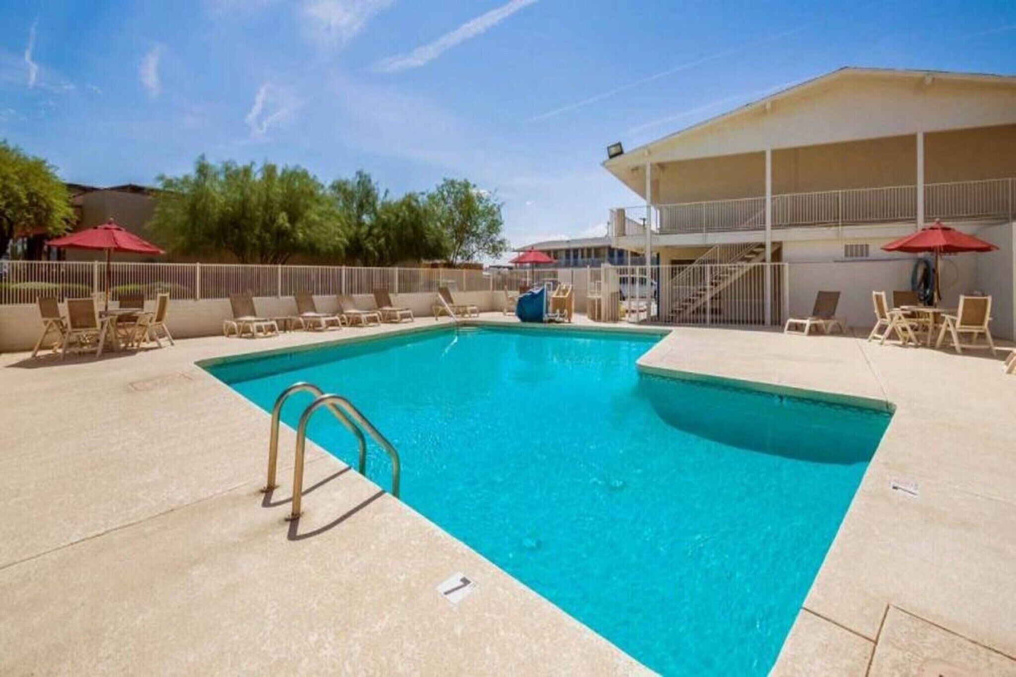 A rectangular swimming pool with a ladder, surrounded by lounge chairs and umbrellas, with a building in the background.