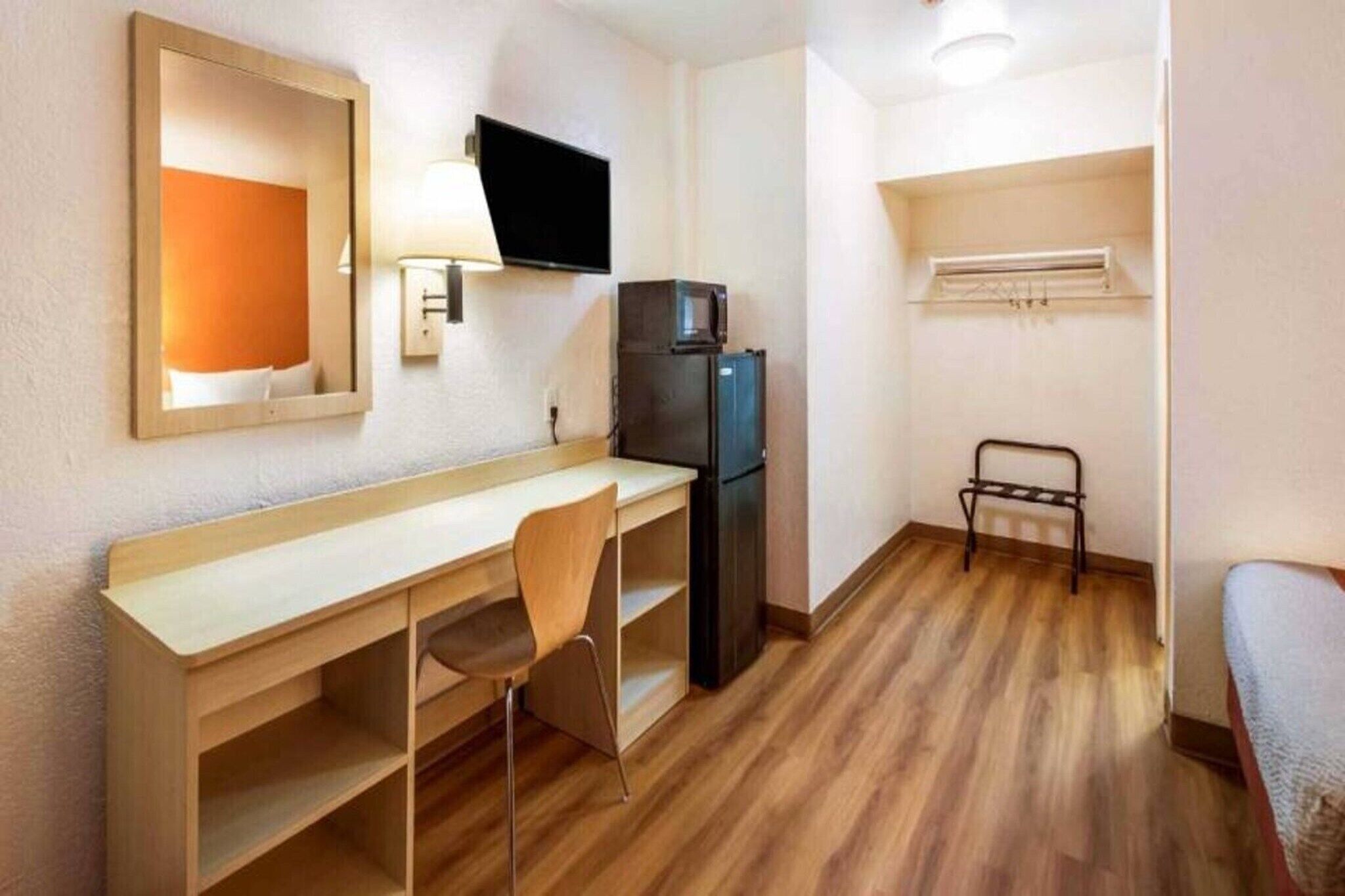 A compact hotel room with a wooden desk, a chair, a black refrigerator, a television mounted on the wall, and a bed with a metal frame.