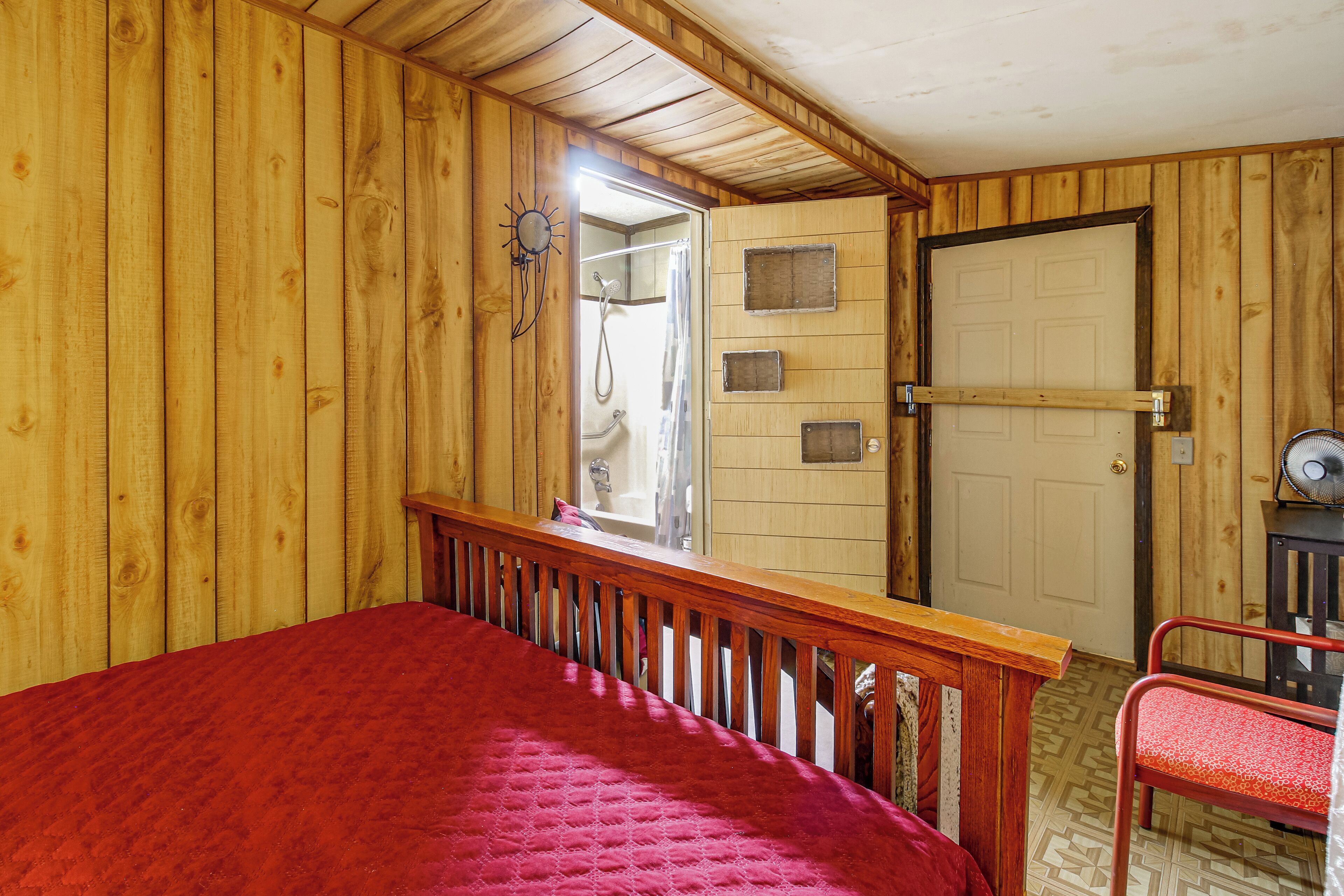 A room with wooden walls, a bed with a red bedspread, a wooden railing, a door with a towel rack, and a fan on a table.