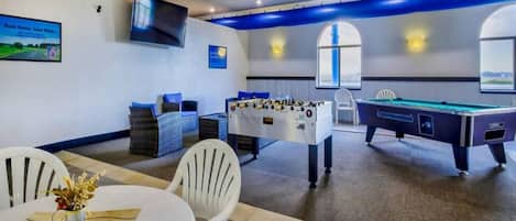 A room with a pool table, foosball table, and a table with chairs and a vase of flowers.