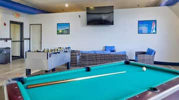 A pool table with a foosball table and seating area in a room with a flat-screen TV mounted on the wall.