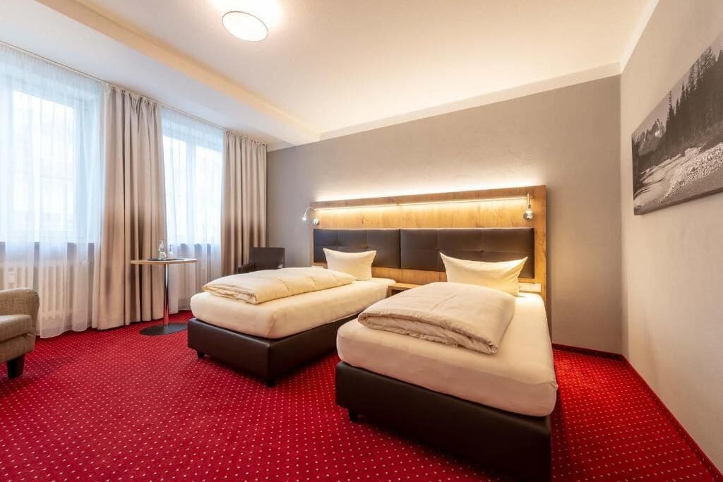 Une chambre d’hôtel avec deux lits, un bureau, une chaise et une grande fenêtre avec des rideaux.