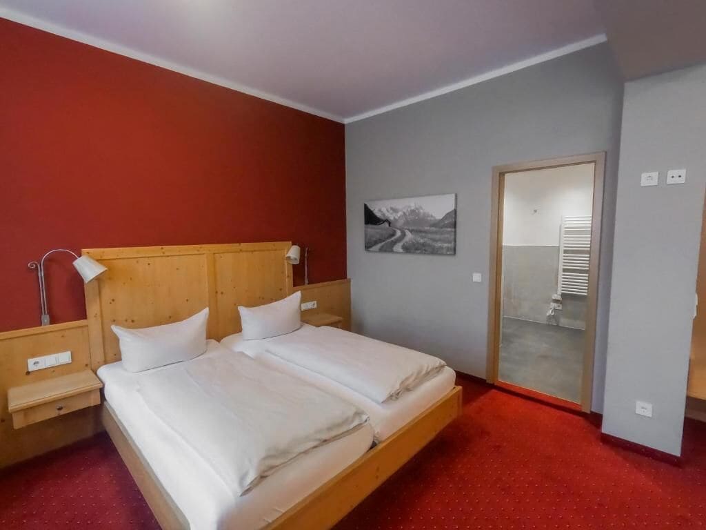 A double bed with a wooden headboard, a bedside table, a wall-mounted lamp, and a picture of a mountainous road.