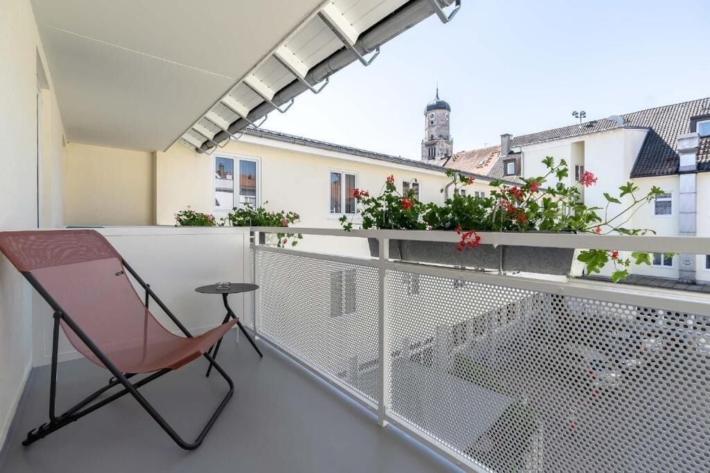 A balcony with a chair and table, a metal railing, and a view of buildings with a tower.