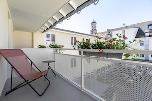 Un balcón con una silla y una mesa, barandilla metálica y vistas a edificios con una torre.