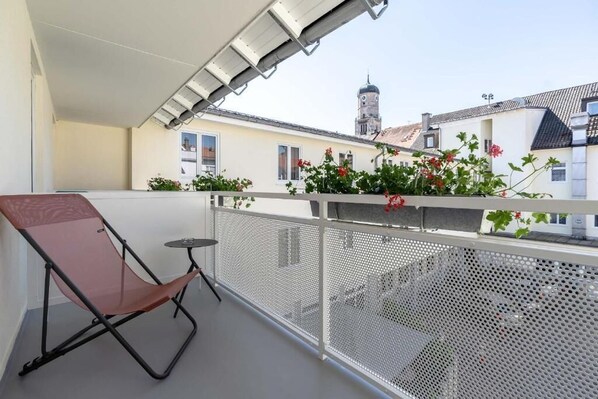 Un balcón con una silla y una mesa, barandilla metálica y vistas a edificios con una torre.