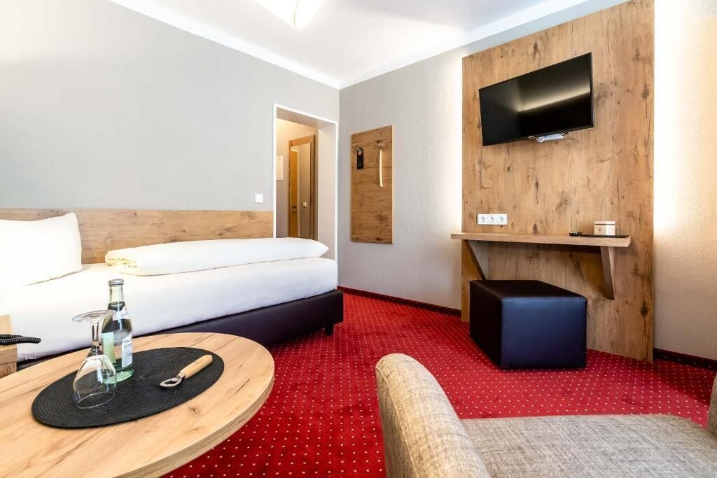 A hotel room with a bed, a TV mounted on a wooden wall, a small table with a bottle and glass, and a red carpeted floor.