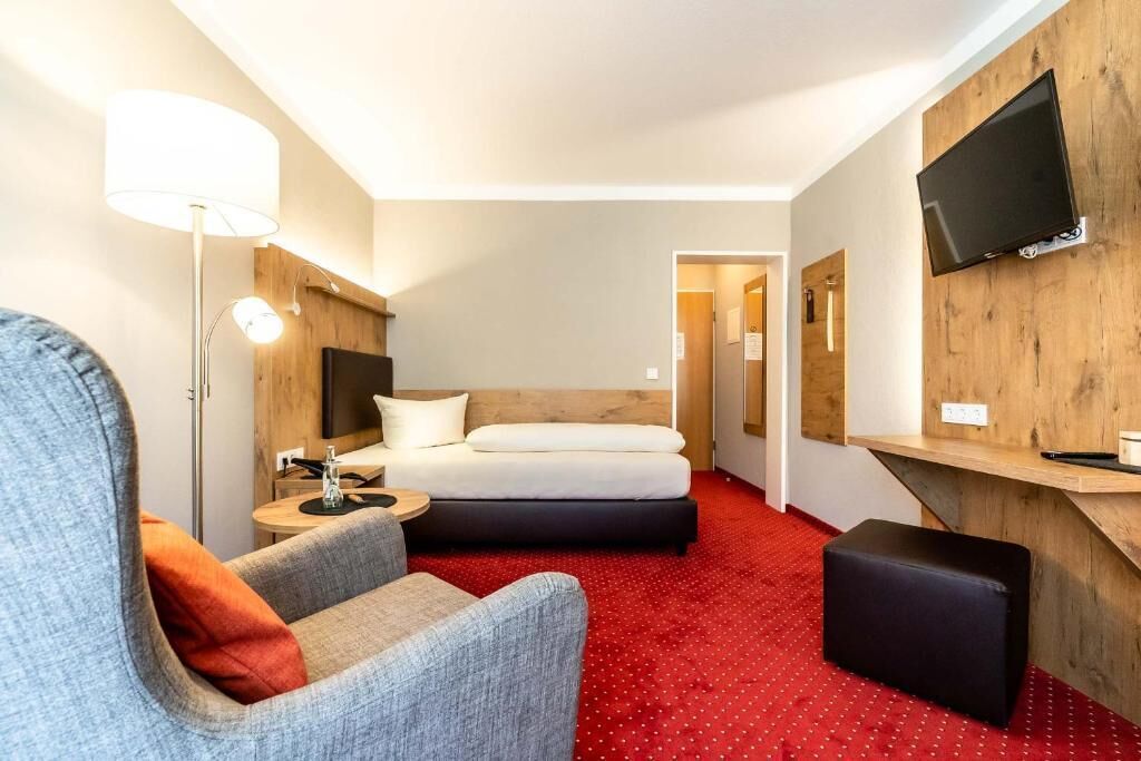 A hotel room with a bed, a desk, a chair, a TV, and a red carpet.